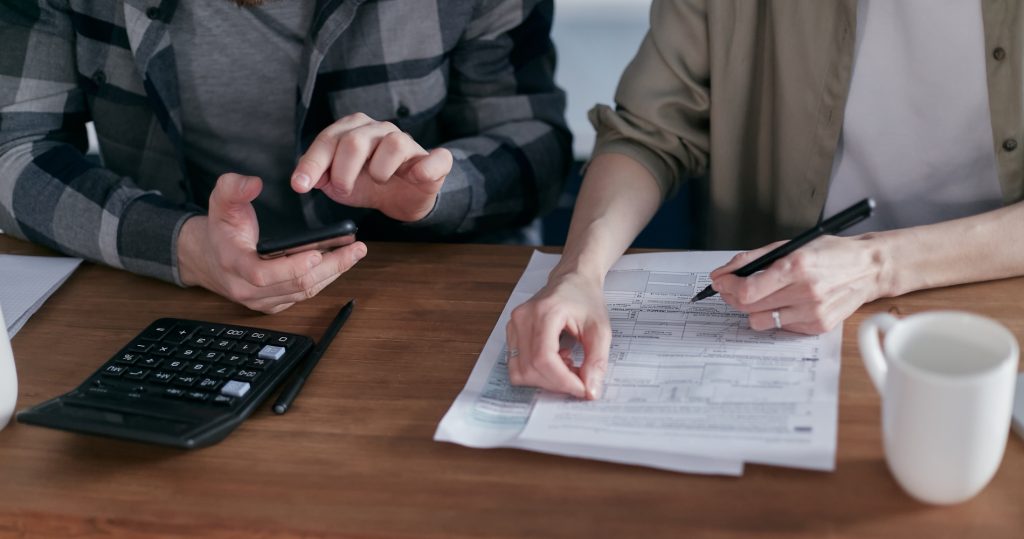 Couple engaged in strategic tax planning at a wooden table, with one person reviewing tax forms using a pen and the other using a smartphone next to a calculator, symbolizing financial preparation for San Antonio homeowners.