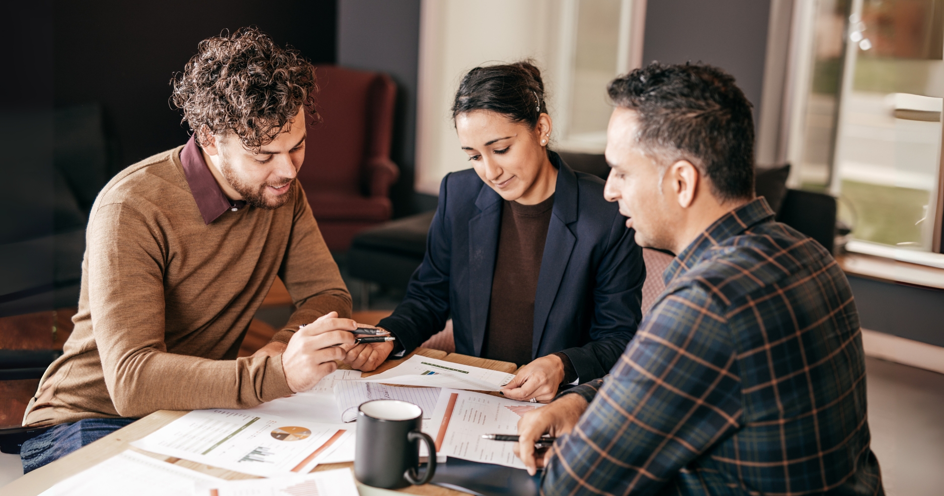 Three people sitting at a table reviewing financial documents together, discussing mortgage or loan options in a professional setting with charts and graphs on paper.