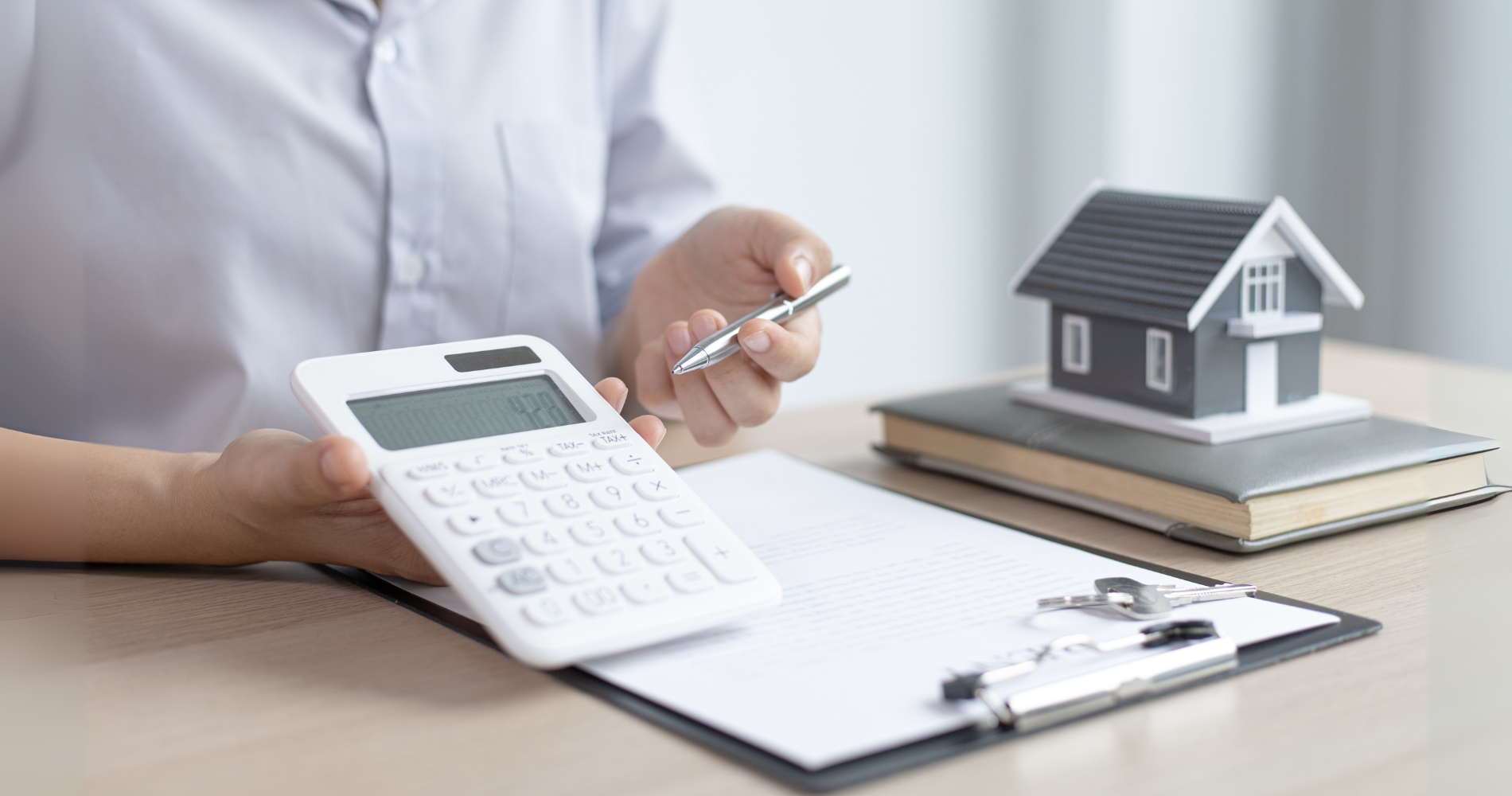 A person is seated at a desk, holding a white calculator in one hand and a silver pen in the other. In front of them on the desk is a clipboard with a document, and next to it, a small model house sits atop a book. The image visually represents the concept of calculating the minimum down payment on a conventional loan.